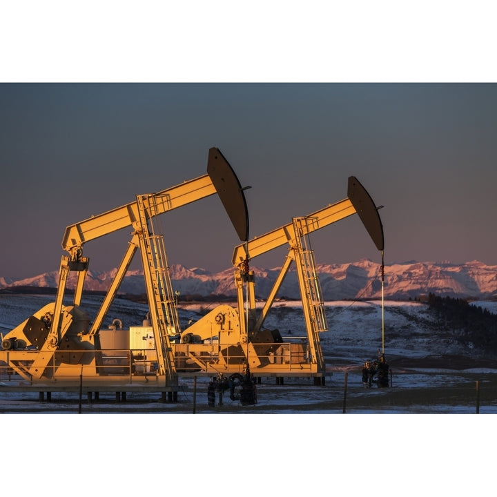 Two pump jacks at sunrise reflecting the warm morning light in a snow covered field and snow covered foothills and mount Image 1