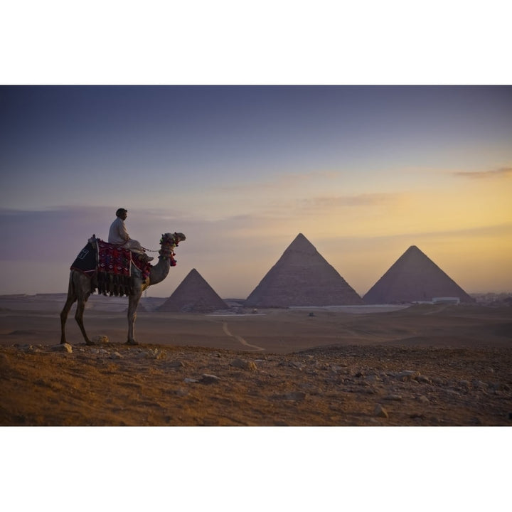 A lone camel and rider stand in front of the setting sun with the great pyramids behind them; Giza Egypt Image 1