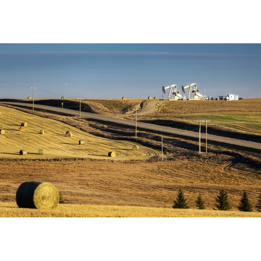 Two pump jacks on rolling hills in the distance along highway with cut fields and hay bales; Alberta Canada Poster Prin Image 1