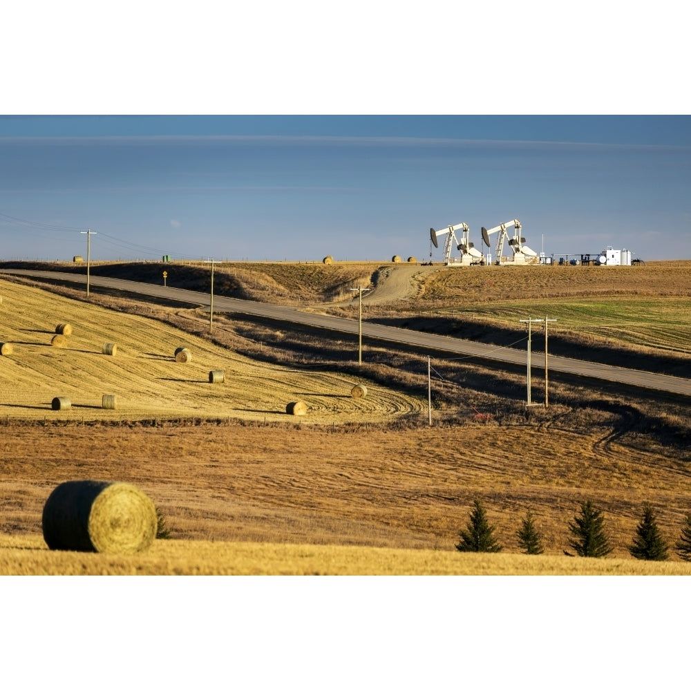 Two pump jacks on rolling hills in the distance along highway with cut fields and hay bales; Alberta Canada Poster Prin Image 2