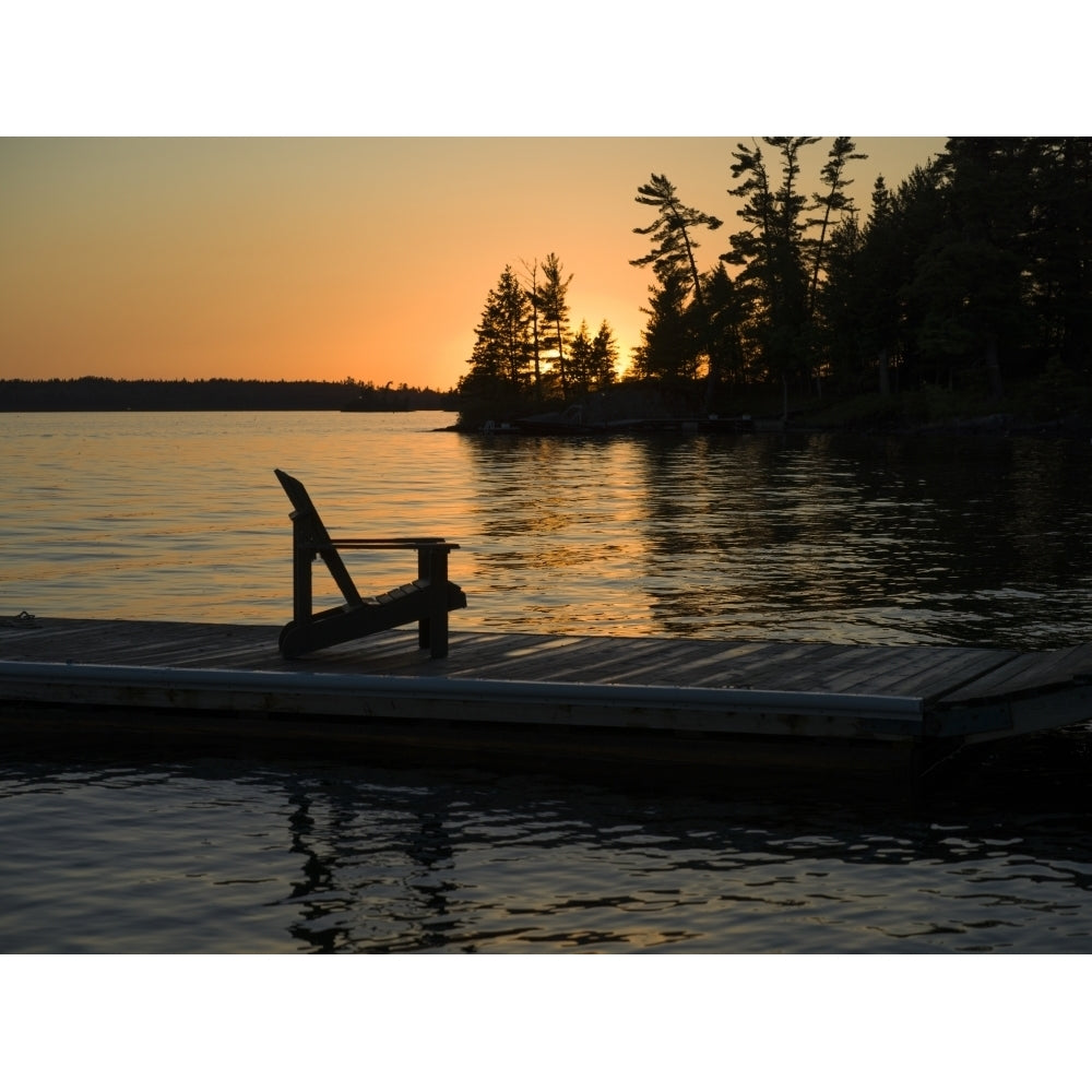 A wooden Adirondack chair sits on a dock on a tranquil lake at sunset; Lake of the Woods Ontario Canada by Keith Levit / Image 1