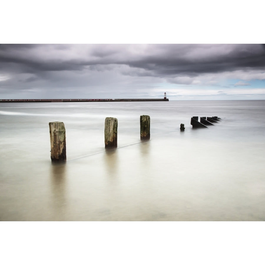 Wooden posts in the ocean with a lighthouse at the end of a pier Poster Print Image 1