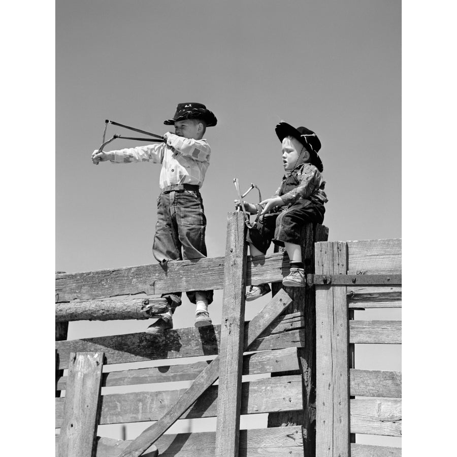 1950s Two Young Boys Dressed As Cowboys Shooting Slingshots On Top Of Wooden Fence Outdoor Print By Vintage Collection Image 1