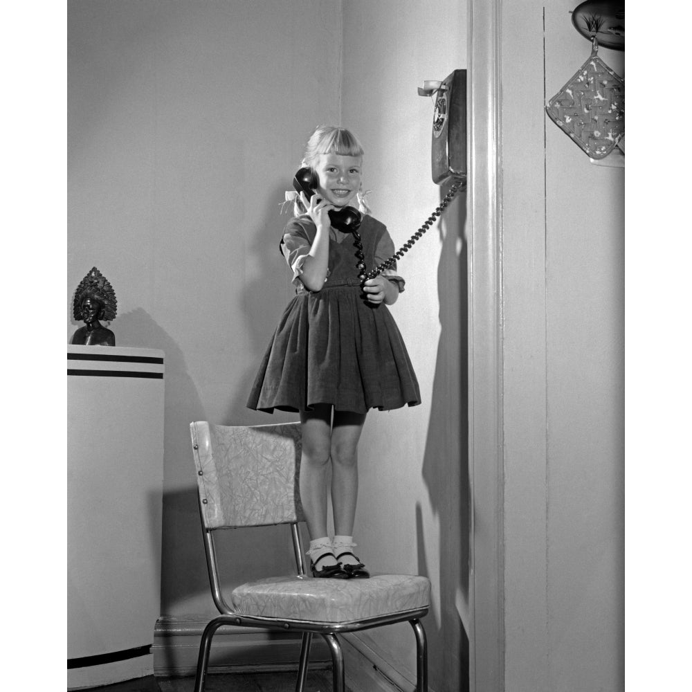 1960s-1950s Young Girl Standing On Kitchen Chair Looking At Camera Talking On Wall Telephone Print By Vintage Collection Image 1