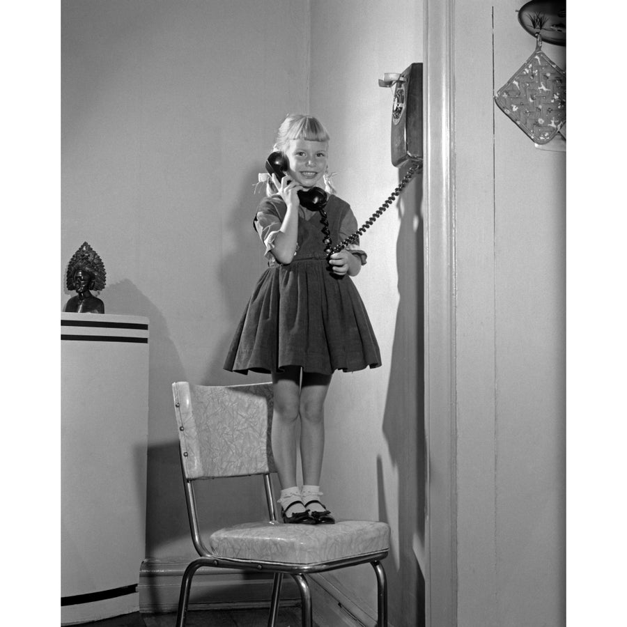 1960s-1950s Young Girl Standing On Kitchen Chair Looking At Camera Talking On Wall Telephone Print By Vintage Collection Image 1